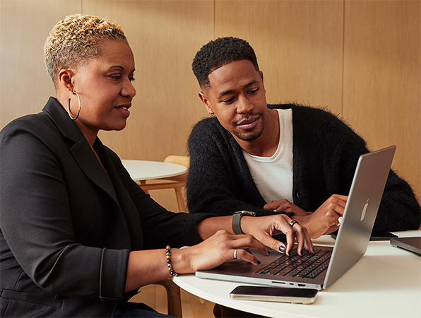 兩位 Apple 同事坐在桌前，使用 MacBook 合作。 