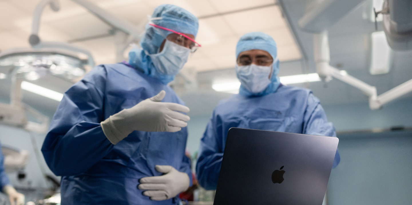 Deux chirurgiens sont dans une salle d’opération et regardent un MacBook; l’un fait un geste de la main tandis que l’autre observe